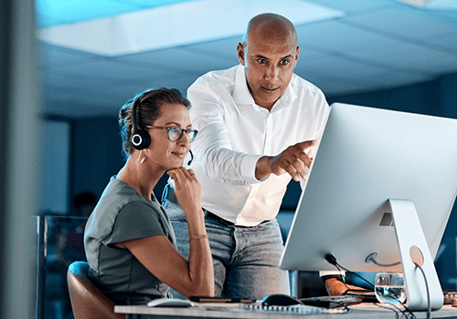 A woman wearing a headset sits at a desk, looking at a computer monitor, while a man standing beside her points at the screen, both appearing focused in a modern office setting.