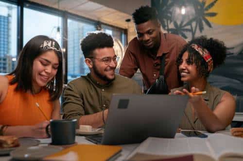 Four young adults sit around a table with a laptop, notebooks, and coffee, smiling and discussing work or studying together in a bright, modern indoor setting.
