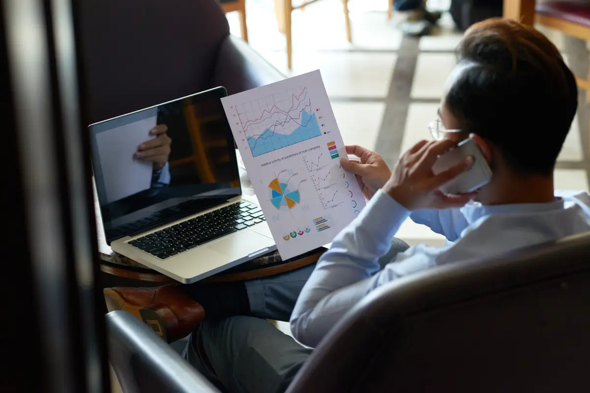 A person sits in a chair holding a paper with charts and graphs, talking on a phone, with an open laptop in front of them on a small table.