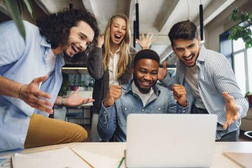 Four young adults excitedly celebrate together while looking at a laptop in a modern office space, showing joyful reactions and raised hands.