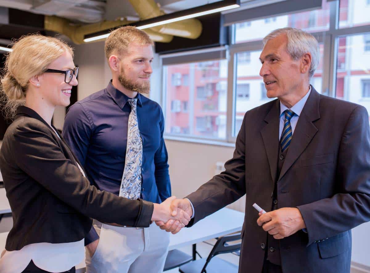 Three people in business attire stand indoors. A woman and an older man are shaking hands and smiling, while a younger man stands between them, watching. Large windows and modern office decor are visible in the background.