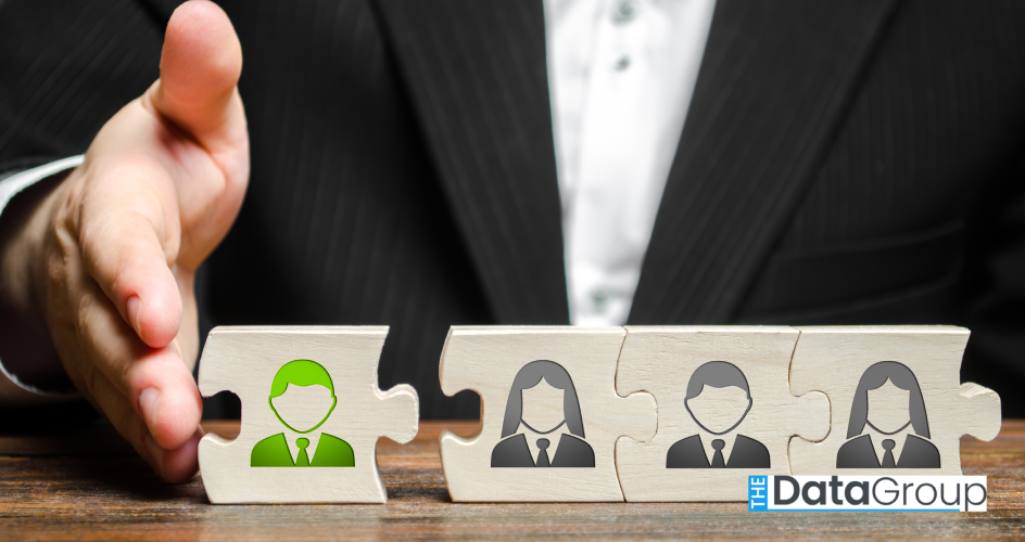 A person wearing a dark jacket and white shirt extends their hand over a wooden table. The hand is above four puzzle pieces. Each piece has an image of a torso with a tie or scarf. One piece is separated from the rest and is colored green. The lighting is bright. The photo is perfect for projects centered on business, teamwork, management, strategy, and office moments.