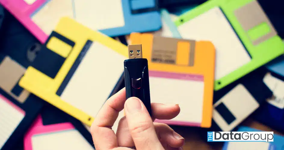 A hand holds a modern USB drive amidst a collection of retro floppy disks of various colors. The shot, taken indoors, is in full daylight, and could be used for projects relating to data storage, technology, retro technology, or computing. The overall mood is organic.