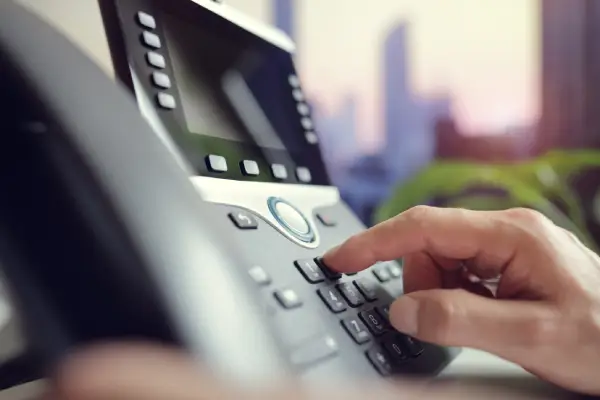Close up of a hand dialing numbers on a modern office telephone. The office setting is suggested by the view of buildings visible in the background. The person's hand presses a button on the gray phone. The scene is shot in natural light, giving the image a sophisticated, business-like feel. This image could be used for projects around workplace communication, office technology, or business strategy