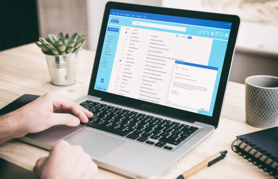 Close up view of hands typing on a laptop keyboard. The laptop is open to an email client program displaying an inbox with various email listings. A notebook, pen, and succulent are also visible on the wooden desk. This photo could be used for projects centered on business, technology, or online communication, emphasizing productivity and information management in a modern office setting. The image can be also used to illustrate tutorials, how-to guides, or articles about email management and internet safety.