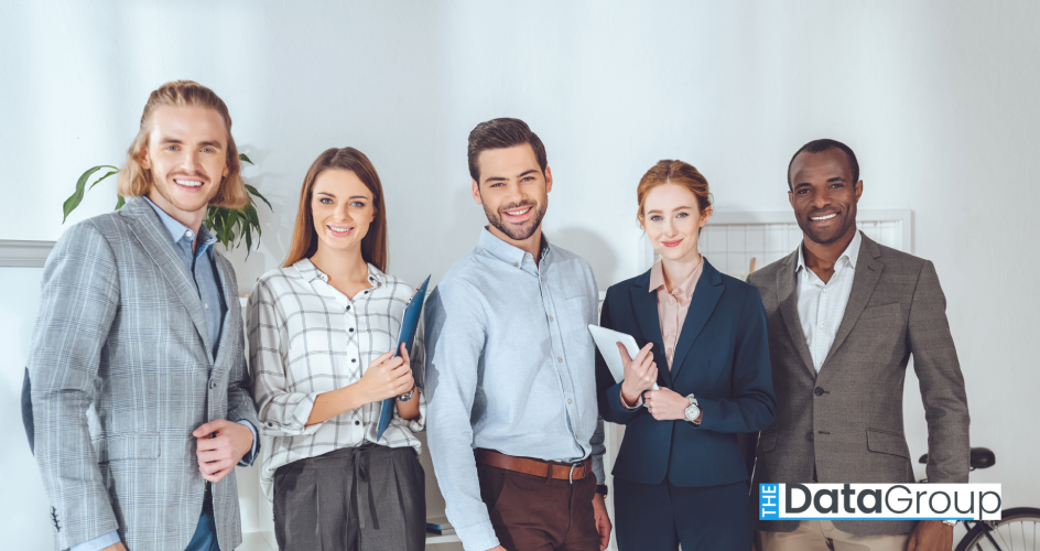 Five business professionals in suits stand outdoors in a city with their arms crossed, smiling confidently.