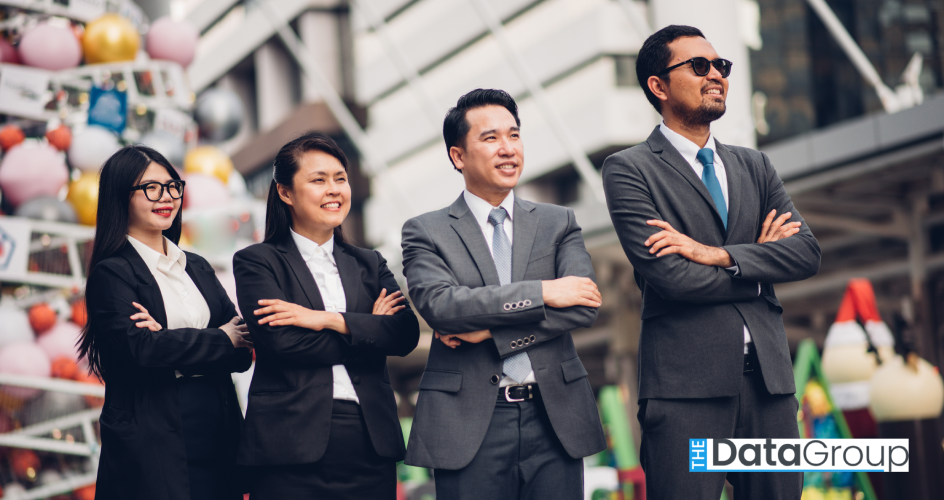 Four business professionals in suits stand outdoors in a city with their arms crossed, smiling confidently.