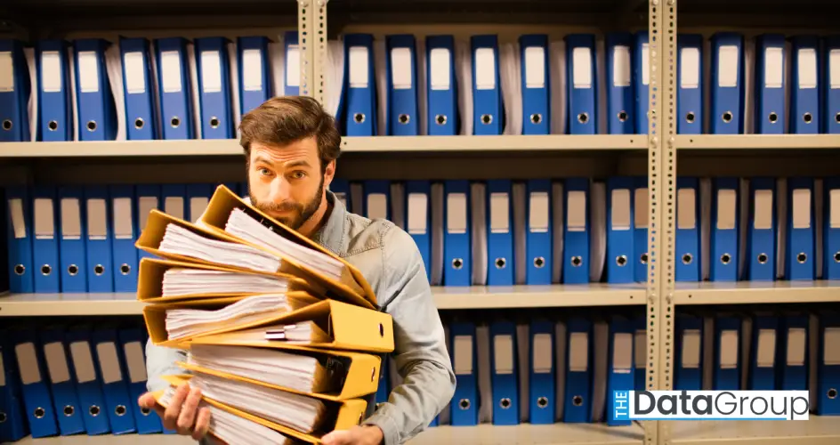 A man with brown hair and a beard, dressed in a denim shirt, carries a large stack of yellow file folders. The photo is shot at waist-up level. Behind him is a metal shelving unit holding numerous blue file binders. The scene is indoors, lit with diffuse, even light. This image could be used for projects about archiving, records management, or the challenges of overwhelming bureaucracy. It could also be used in articles about the need for automation and efficient systems.