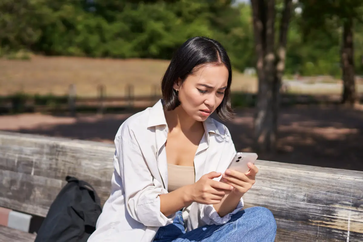 A woman sits on a wooden bench outdoors, looking at her smartphone with a concerned expression. She is wearing a light shirt and jeans, with trees and a blurred park background behind her.