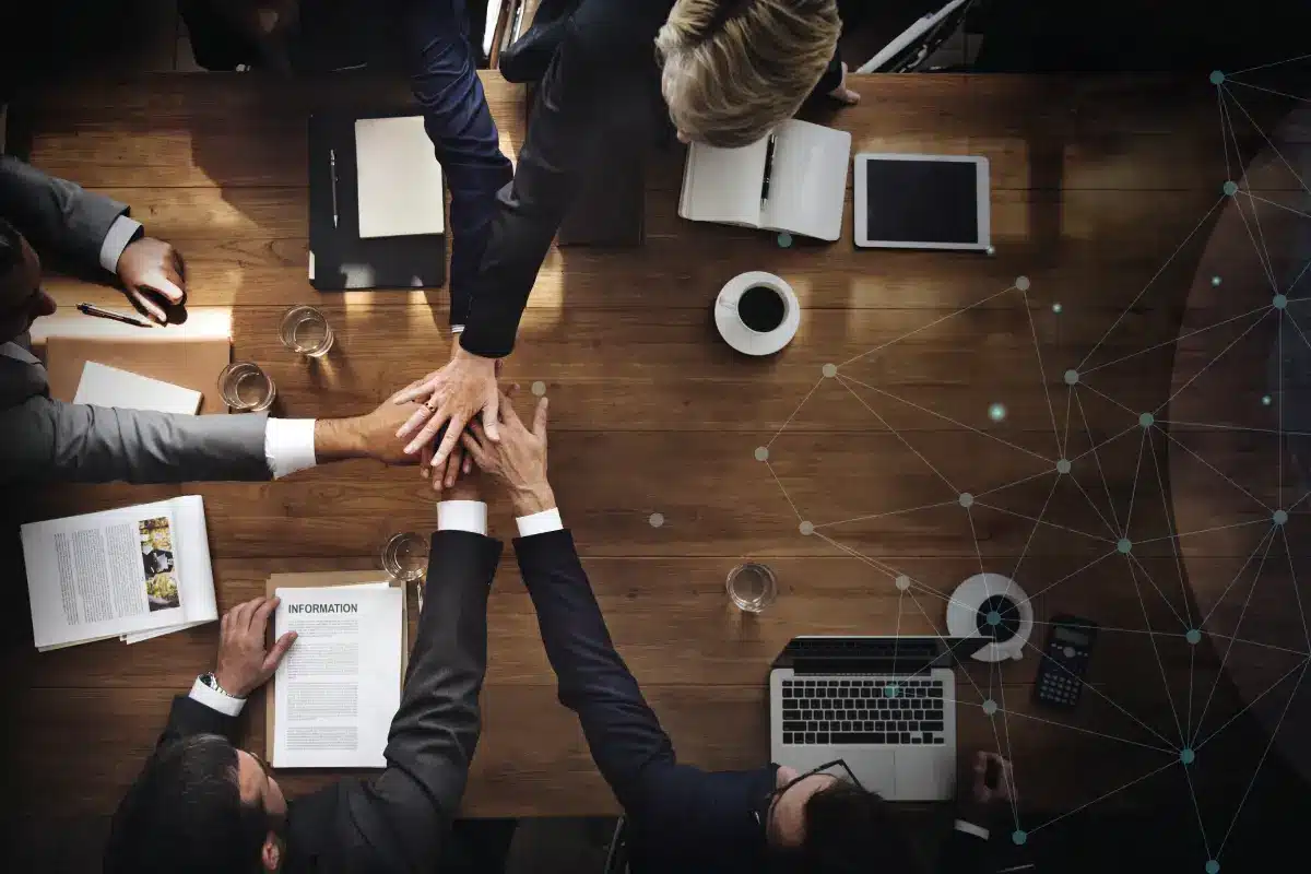 A group of businesspeople sitting around a wooden table in an office, placing their hands together in the center as a sign of teamwork. Laptops, documents, coffee, and office supplies are visible on the table.