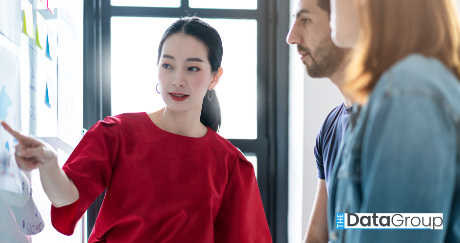 Woman in a bright red blouse points to documents on a wall while two coworkers stand beside her in a sunlit office. The group appears to be reviewing ideas or project materials during a team discussion.
