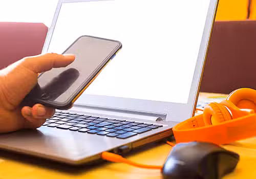 A person holds a smartphone near an open laptop with a blank white screen. Orange headphones and a black mouse are on the table beside the laptop.
