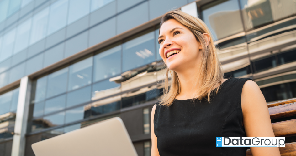 Smiling woman in a sleeveless black dress sits outdoors with an open laptop in front of a glass office building. The scene suggests remote work or a business meeting in an urban setting.