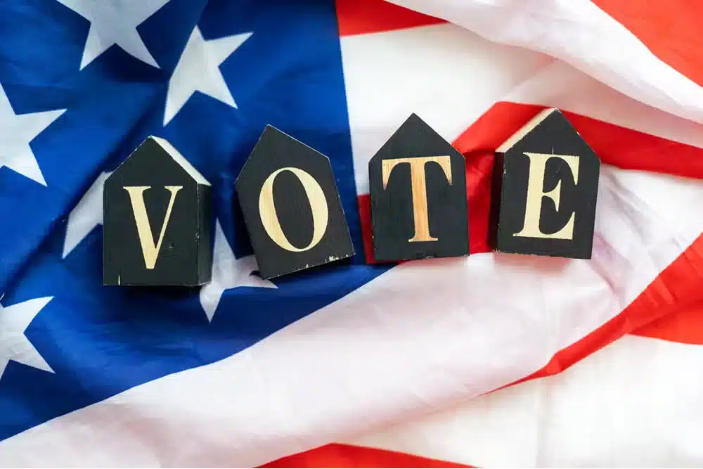 Four black blocks with gold letters spelling VOTE are arranged on a crumpled section of an American flag with visible stars and stripes, symbolizing the importance of joining mailing lists to stay informed about election updates.