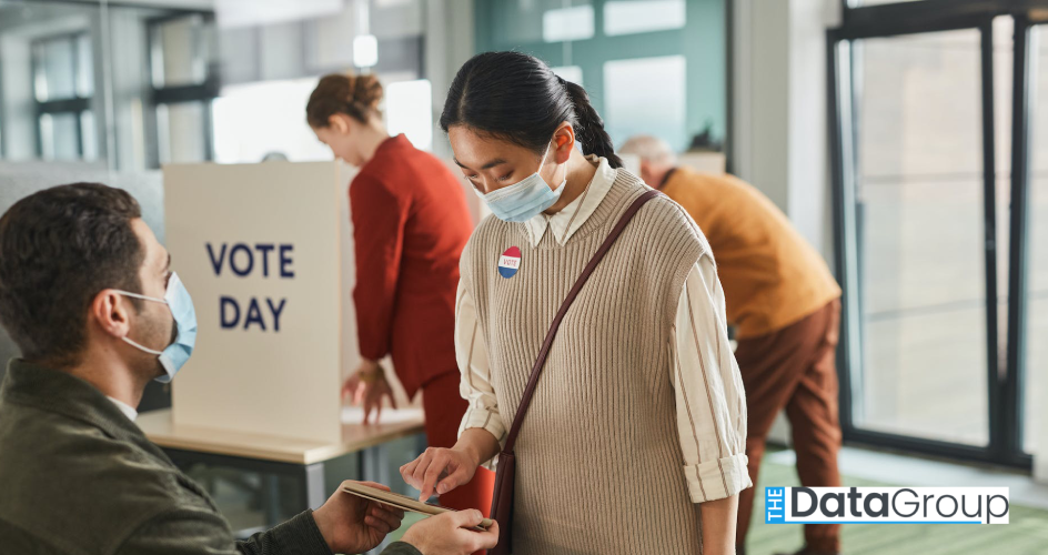 Masked voter checks in at a polling station by pointing to a tablet held by a poll worker. A booth behind them reads "VOTE DAY" while other voters cast ballots in the background.
