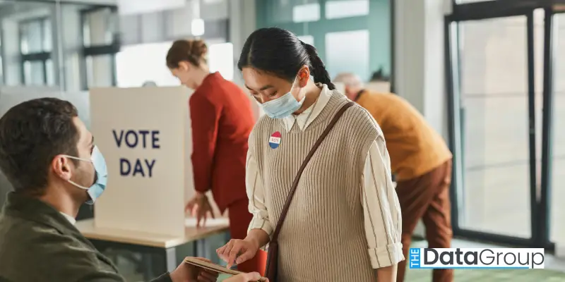 A woman wearing a face mask and an I Voted sticker checks in at a polling station on election day, while other people vote in the background.