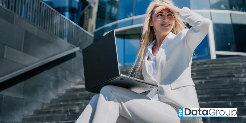 A woman in a white suit sits on outdoor steps with a laptop on her lap, smiling and shielding her eyes from the sun. The Data Group logo appears in the bottom right corner. Modern glass building in the background.