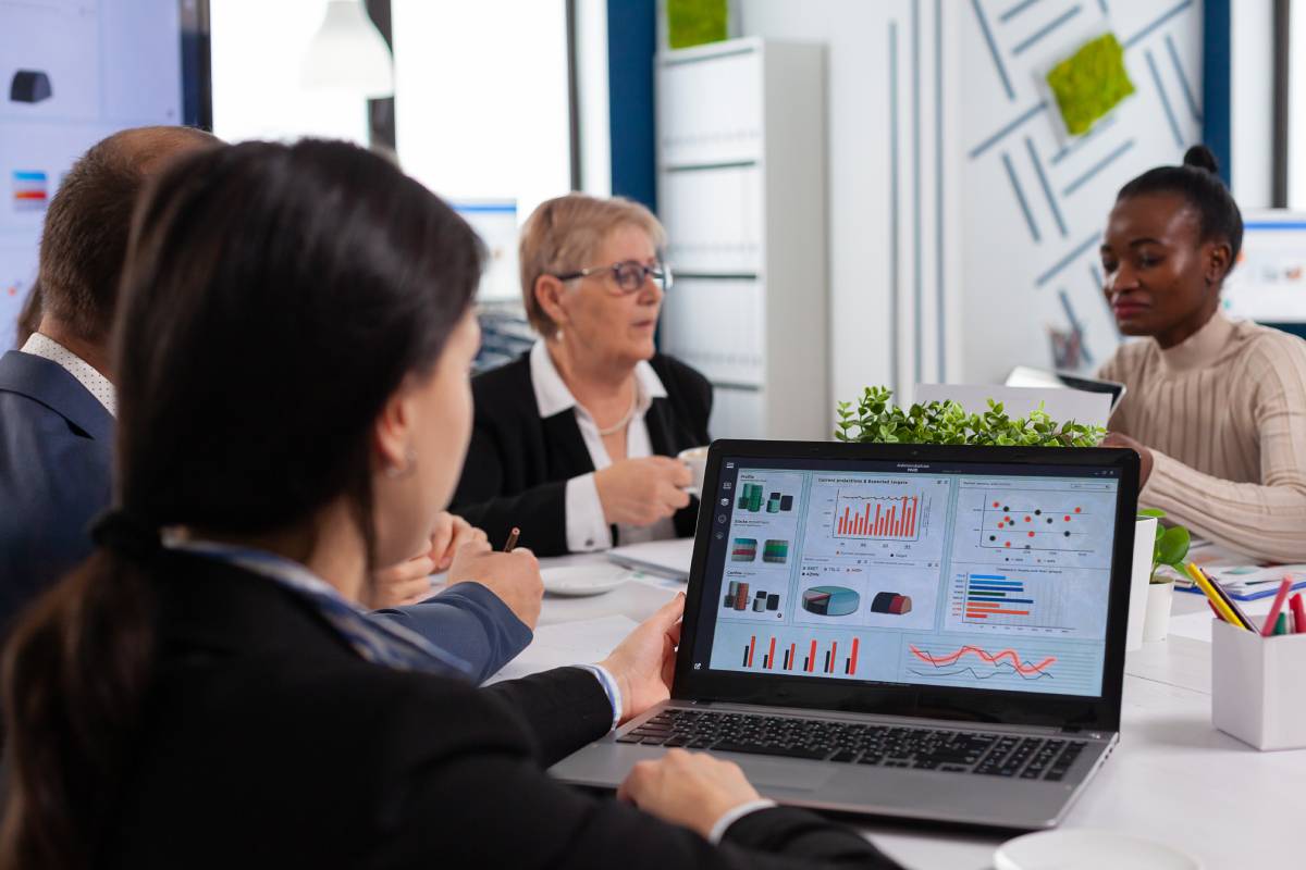 A group of business professionals sits at a conference table. One person displays charts and graphs on a laptop screen, illustrating how using data append can improve marketing engagement, while others listen and discuss in a modern office setting.