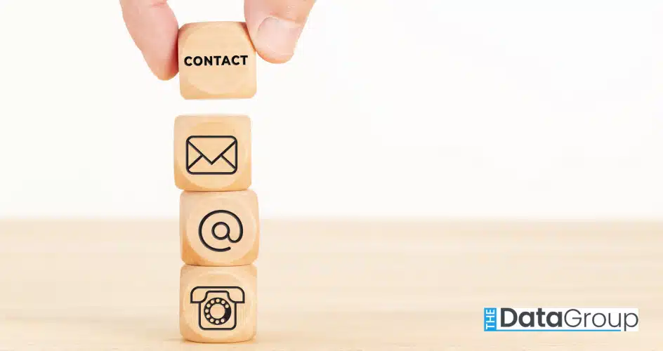 This close-up shot features a hand carefully placing a wooden block labeled "Contact" atop a stack of similarly styled blocks. Each block in the stack showcases a different contact method, including icons for email, at symbol, and a telephone, against a crisp white background. The composition creates a clean, minimalist aesthetic, ideal for business and technology projects, website design, or informational graphics. This image evokes professionalism and efficiency and conveys messages related to connection and accessibility.